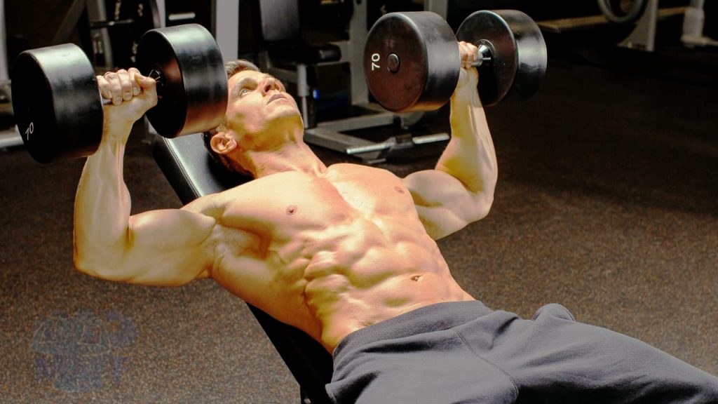 Buff shirtless man performing incline dumbbell bench press in a gym.